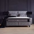 Babylon Boxspringbett in Samt Grau als Schlafzimmer-Szene mit gepolstertem Kopfteil, Matratze und Topper vor dunkler Wand