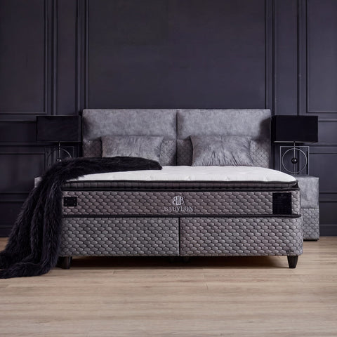 Babylon Boxspringbett in Samt Grau als Schlafzimmer-Szene mit gepolstertem Kopfteil, Matratze und Topper vor dunkler Wand