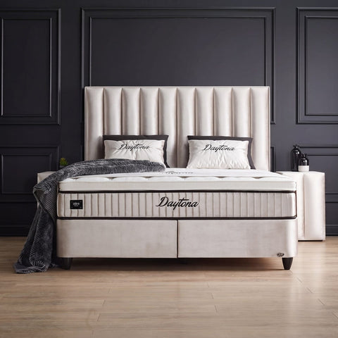 Daytona Boxspringbett in Samt Hell Beige, Frontansicht mit vertikal gestepptem Kopfteil, Matratze und Topper vor dunkler Wand