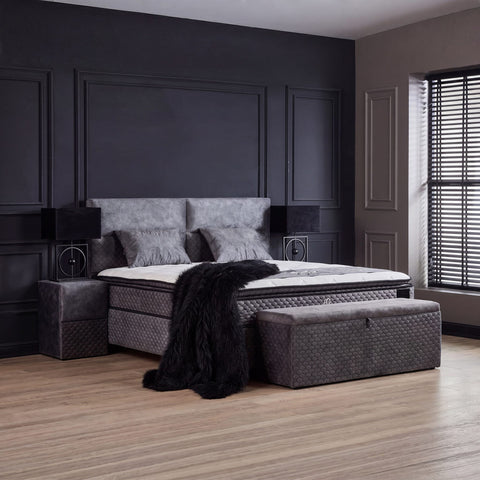Babylon Boxspringbett in Grau mit Nachttischen und Stauraum-Truhe im dunklen Schlafzimmer arrangiert