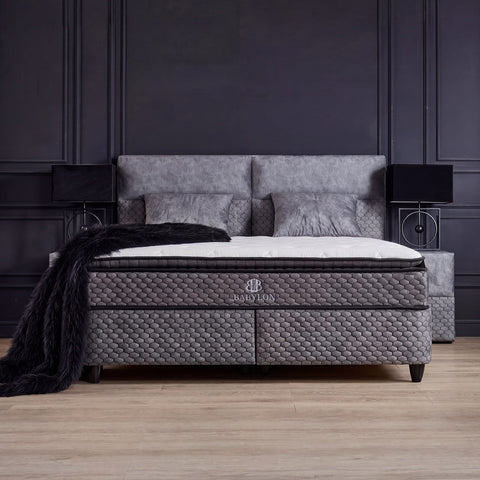Babylon Boxspringbett in Samt Grau als Schlafzimmer-Szene mit gepolstertem Kopfteil, Matratze und Topper vor dunkler Wand