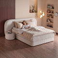 Brooklyn Boxspringbett Samt Beige mit Frau auf der Liegefläche, hoher Liegekomfort von Matratze und Topper im warmen Schlafzimmer