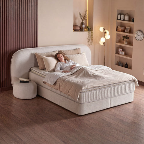Brooklyn Boxspringbett Samt Beige mit Frau auf der Liegefläche, hoher Liegekomfort von Matratze und Topper im warmen Schlafzimmer